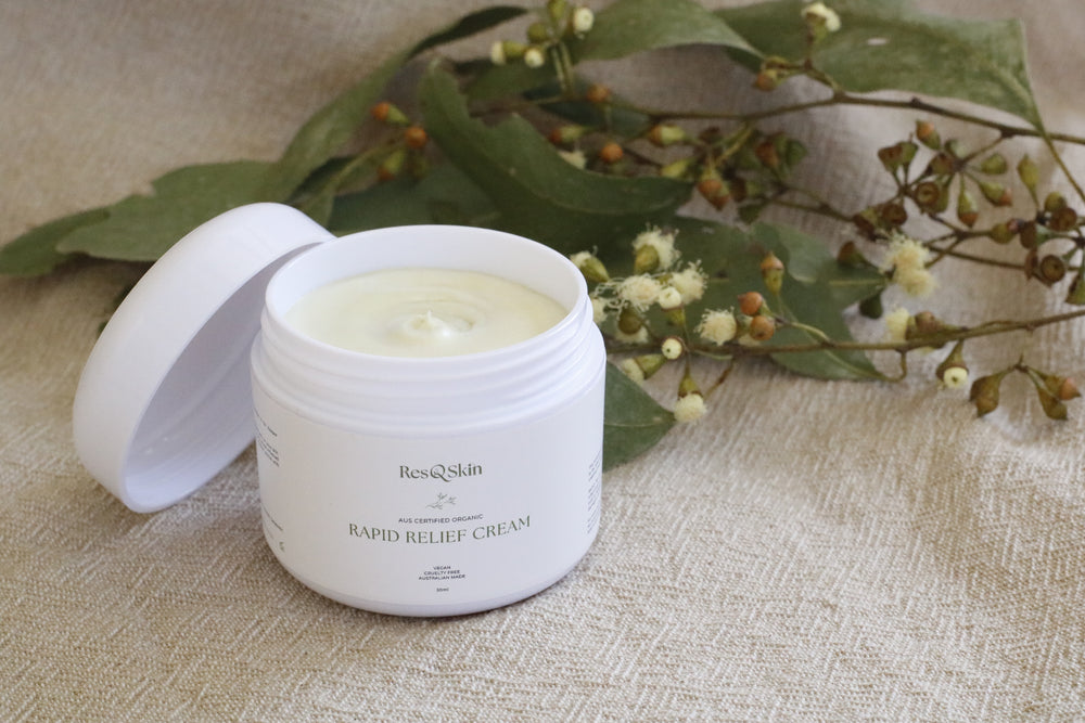 A 50g tub of Rapid Relief Cream with ingredients and essentials oils in the background. The tub has the lid off showing the creamy thick white relief cream